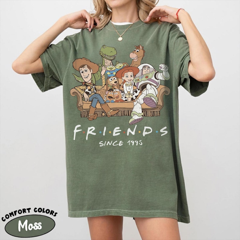 May include: A moss green short-sleeve t-shirt featuring a graphic of Toy Story characters sitting on a couch with the text "FRIENDS SINCE 1995" printed below. The characters include Woody, Buzz Lightyear, Jessie, Rex, Bullseye, and others.