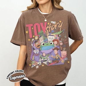 Op de afbeelding: Een bruin T-shirt met korte mouwen en een grote grafische print met de tekst TOY story. Het ontwerp bevat diverse geanimeerde figuren zoals een cowboy, een space ranger, een aardappelfiguur, een dinosaurus en een varken rondom een paarse tas.