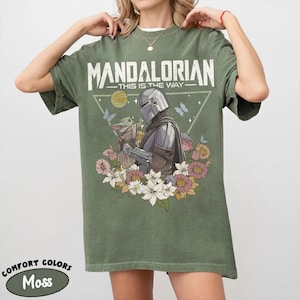 May include: Moss green t-shirt featuring a graphic of The Mandalorian and Grogu surrounded by flowers and butterflies. The text "MANDALORIAN THIS IS THE WAY" is printed across the top. The shirt is a Comfort Colors brand.