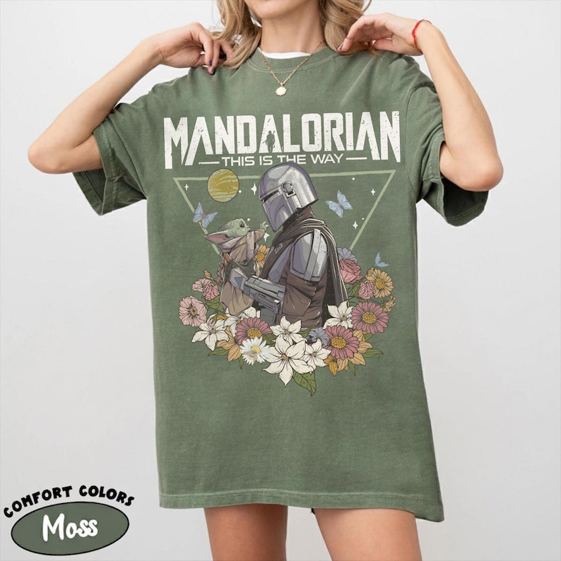 May include: Moss green t-shirt featuring a graphic of The Mandalorian and Grogu surrounded by flowers and butterflies. The text "MANDALORIAN THIS IS THE WAY" is printed across the top. The shirt is a Comfort Colors brand.