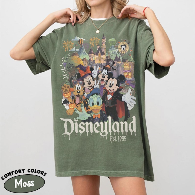 May include: A moss green short-sleeve t-shirt featuring a graphic print of Disney characters in Halloween costumes, including Mickey Mouse, Minnie Mouse, Goofy, Pluto, and Donald Duck in front of a castle. The text reads Disneyland Est. 1955.