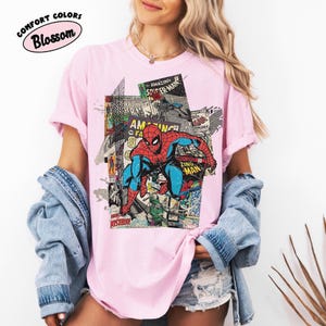 Pode incluir: T-shirt rosa claro com um gr&aacute;fico de banda desenhada do Homem-Aranha. O design incorpora pain&eacute;is de banda desenhada e elementos de texto. A t-shirt tem o texto "Comfort Colors Blossom" num oval rosa.