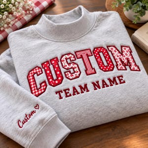 May include: A light gray mock neck sweatshirt featuring the word CUSTOM in large, red, patterned varsity-style letters, with the text TEAM NAME below it. The sleeve includes the word Custom and a small heart embroidered in red thread.