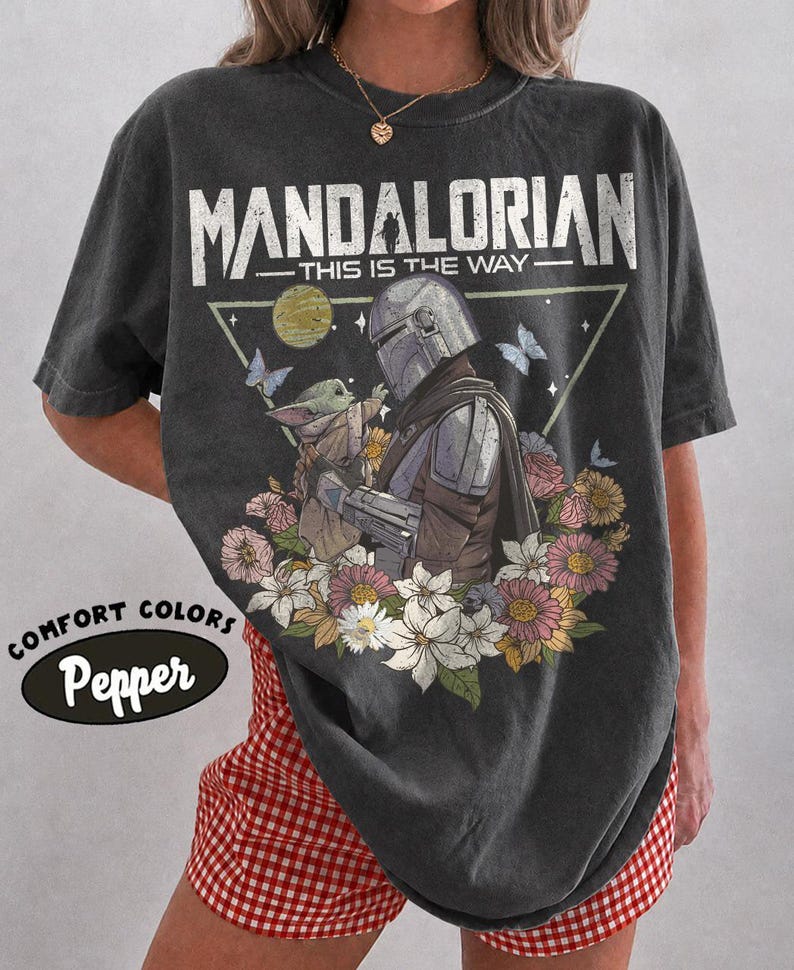 May include: Dark gray t-shirt featuring a graphic of The Mandalorian and Grogu surrounded by flowers and butterflies. The text "MANDALORIAN THIS IS THE WAY" is printed above the image. The shirt has a vintage look and the text "COMFORT COLORS Pepper" is printed on the bottom left.