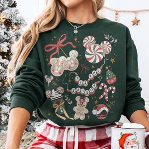 May include: A dark green sweatshirt featuring a festive design with gingerbread men, candy canes, and the phrase "Tis the Season." The design includes red, white, and gold accents, creating a holiday theme. The sweatshirt is a crew neck style.