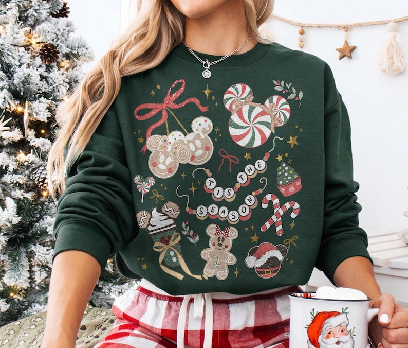 May include: A dark green sweatshirt featuring a festive design with gingerbread men, candy canes, and the phrase "Tis the Season." The design includes red, white, and gold accents, creating a holiday theme. The sweatshirt is a crew neck style.