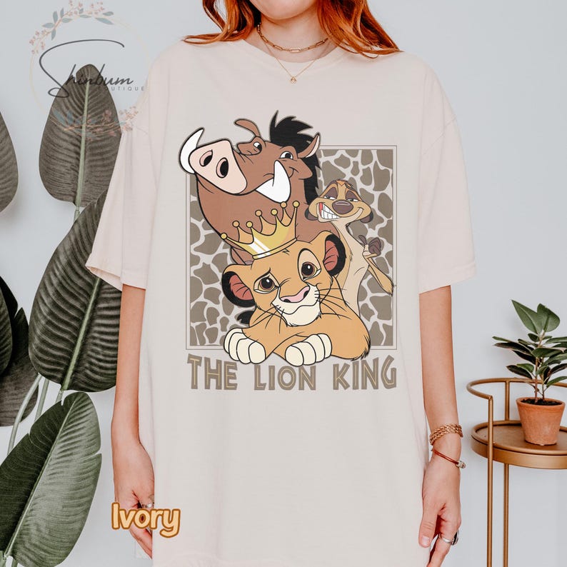 May include: Ivory-colored t-shirt featuring a graphic of Simba wearing a crown, Pumbaa, and Timon from The Lion King. The text "THE LION KING" is below the characters. The design is set against a brown and tan pattern.