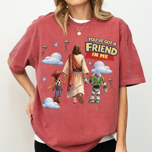 May include: Red t-shirt with a graphic of Jesus walking with Woody and Buzz Lightyear. The text "YOU'VE GOT A FRIEND IN ME" is displayed in yellow and blue. The design includes clouds, stars, and parachuting figures.