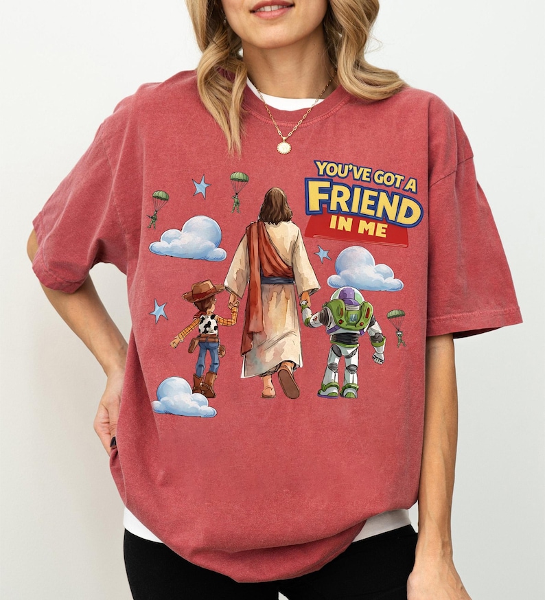 May include: Red t-shirt with a graphic of Jesus walking with Woody and Buzz Lightyear. The text "YOU'VE GOT A FRIEND IN ME" is displayed in yellow and blue. The design includes clouds, stars, and parachuting figures.