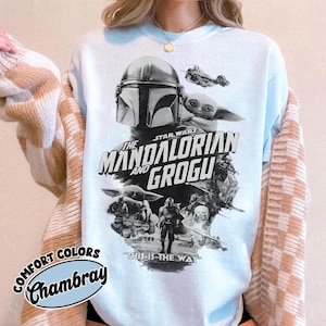 Op de afbeelding: Lichtblauw sweatshirt met een afbeelding uit de Star Wars-serie, "The Mandalorian and Grogu". Het ontwerp bevat de helm van de Mandalorian, Grogu en de zin "This is the Way". Het sweatshirt heeft een "Comfort Colors Chambray" label.