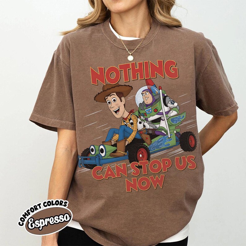 May include: A brown short-sleeve t-shirt featuring a graphic of Woody and Buzz Lightyear riding a green race car. The text "NOTHING CAN STOP US NOW" is printed in red above and below the characters. The shirt has a crew neckline and a relaxed fit.