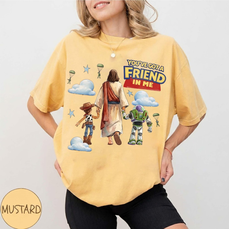 May include: Mustard yellow t-shirt with a graphic of Jesus, Woody, and Buzz Lightyear. The text "YOU'VE GOT A FRIEND IN ME" is above the characters. The design includes clouds, stars, and parachutes. The t-shirt is a comfortable fit.