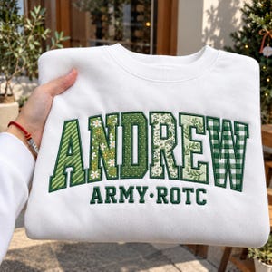 May include: A white crewneck sweatshirt featuring the name ANDREW in large, block-style letters with varied green patterned fabric appliques. Below the name, the text ARMY ROTC is embroidered in solid green thread on the front of the garment.