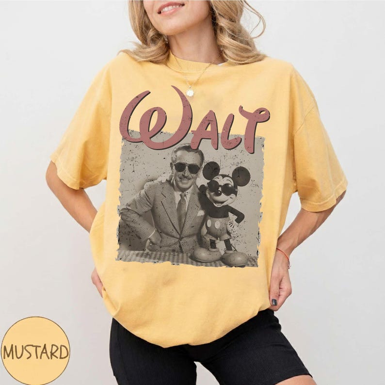 May include: Mustard yellow t-shirt featuring a vintage graphic of Walt Disney with Mickey Mouse. The graphic is in a rectangular frame with the word "Walt" in a burgundy script across the top. The shirt is a relaxed fit.