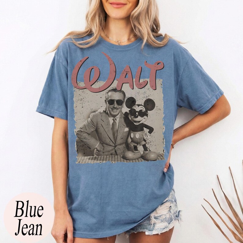 May include: Blue jean colored t-shirt featuring a vintage graphic of Walt Disney and Mickey Mouse wearing sunglasses. The word "WALT" is printed above the image in a distressed pink font. The words "Blue Jean" are printed in white at the bottom.