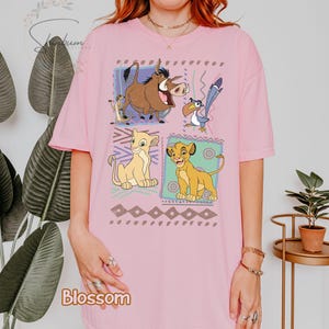 May include: Pink t-shirt featuring a colorful graphic of characters from The Lion King. The design includes Simba, Nala, Timon, Pumbaa, and Zazu, set against a geometric pattern. The word "Blossom" is printed at the bottom.