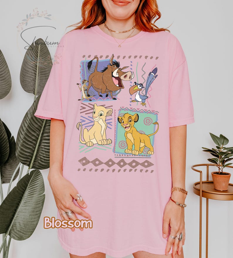 May include: Pink t-shirt featuring a colorful graphic of characters from The Lion King. The design includes Simba, Nala, Timon, Pumbaa, and Zazu, set against a geometric pattern. The word "Blossom" is printed at the bottom.