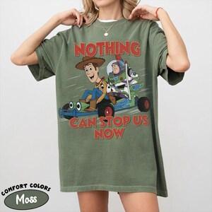 May include: An olive green short-sleeve t-shirt featuring a graphic of Woody and Buzz Lightyear riding a go-kart. The text "NOTHING CAN STOP US NOW" is printed in red above and below the characters. The shirt has a crew neckline.