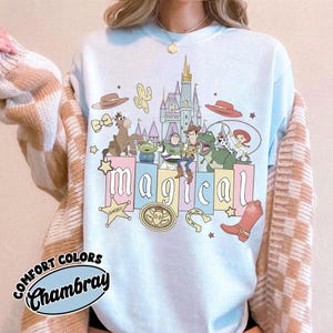 Puede incluir: Sudadera azul claro con un gr&aacute;fico colorido de personajes de dibujos animados, un castillo y la palabra "m&aacute;gico". Gr&aacute;ficos adicionales incluyen un sombrero de vaquero, un cactus y una placa de sheriff. La sudadera tiene la frase "Comfort Colors Chambray".
