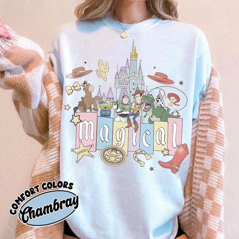 Puede incluir: Sudadera azul claro con un gr&aacute;fico colorido de personajes de dibujos animados, un castillo y la palabra "m&aacute;gico". Gr&aacute;ficos adicionales incluyen un sombrero de vaquero, un cactus y una placa de sheriff. La sudadera tiene la frase "Comfort Colors Chambray".