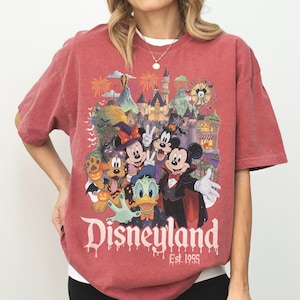 May include: A faded red short-sleeve t-shirt featuring a graphic of Disney characters in Halloween costumes in front of a castle. The text reads Disneyland Est. 1955. The characters include Mickey Mouse, Minnie Mouse, Donald Duck, Goofy, and Pluto.