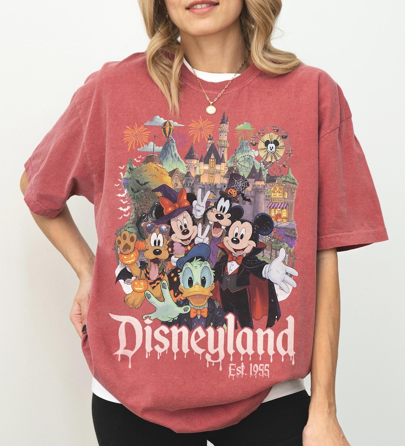 May include: A faded red short-sleeve t-shirt featuring a graphic of Disney characters in Halloween costumes in front of a castle. The text reads Disneyland Est. 1955. The characters include Mickey Mouse, Minnie Mouse, Donald Duck, Goofy, and Pluto.