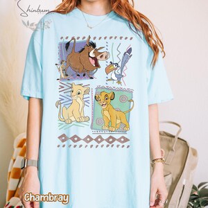 May include: Light blue t-shirt featuring a colorful graphic of characters from The Lion King. The design includes Simba, Timon, Pumbaa, Zazu, and Nala, set against a geometric background. The word "Chambray" is printed on the shirt.