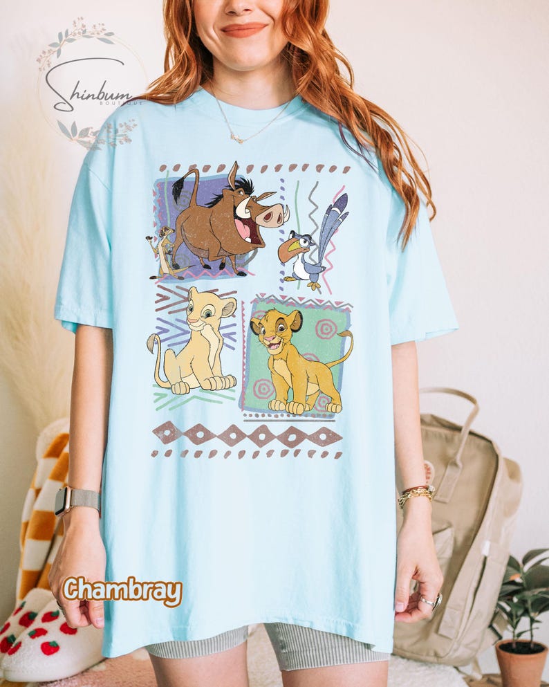 May include: Light blue t-shirt featuring a colorful graphic of characters from The Lion King. The design includes Simba, Timon, Pumbaa, Zazu, and Nala, set against a geometric background. The word "Chambray" is printed on the shirt.