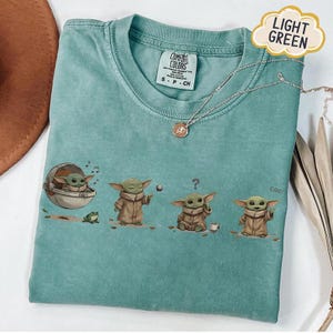 Puede incluir: Camiseta verde claro Comfort Colors con un gr&aacute;fico de un personaje con una t&uacute;nica marr&oacute;n en varias poses. Un collar plateado con un colgante est&aacute; en la camiseta. La camiseta tiene una etiqueta que dice "Comfort Colors".