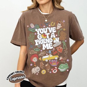 Op de afbeelding: Een bruin t-shirt met korte mouwen en een grafische print met de tekst "YOU'VE GOT A FRIEND IN ME", omringd door diverse kleurrijke illustraties van stripfiguren, een gele vrachtwagen, een cactus en sterren in een collage-stijl.