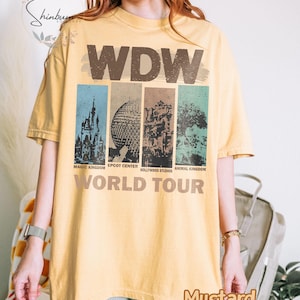 May include: A yellow t-shirt with a vintage-style graphic of the Walt Disney World theme parks. The graphic includes the text "WDW World Tour" and images of Magic Kingdom, Epcot Center, Hollywood Studios, and Animal Kingdom.
