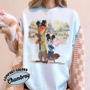 Puede incluir: Sudadera azul claro Comfort Colors Chambray con una ilustraci&oacute;n de dibujos animados de Nick Wilde y Judy Hopps de Zootopia. El dise&ntilde;o incluye una escena de parque con un castillo, carritos de comida y un sol. La sudadera tiene cuello redondo y mangas largas.