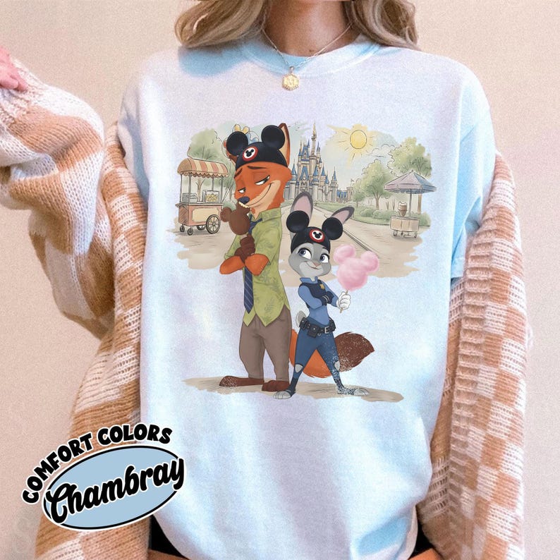 Puede incluir: Sudadera azul claro Comfort Colors Chambray con una ilustraci&oacute;n de dibujos animados de Nick Wilde y Judy Hopps de Zootopia. El dise&ntilde;o incluye una escena de parque con un castillo, carritos de comida y un sol. La sudadera tiene cuello redondo y mangas largas.