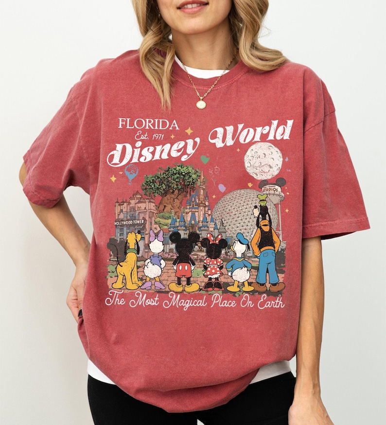 May include: Red t-shirt with a Disney World graphic. The design includes the text "Florida Disney World Est. 1971" with Disney characters and park landmarks. The phrase "The Most Magical Place On Earth" is also included.
