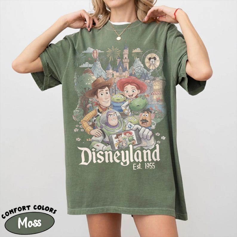 May include: A moss green t-shirt featuring a graphic print of Toy Story characters including Woody, Buzz Lightyear, Jessie, Rex, and Mr. Potato Head taking a selfie in front of a Disneyland castle. The text reads Disneyland Est. 1955.
