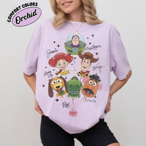 Puede incluir: Una camiseta de color lila claro con un gr&aacute;fico de los personajes de Toy Story: Buzz Lightyear, Woody, Jessie, Slinky Dog, Rex y Mr. Potato. La camiseta tiene el texto "Comfort Colors Orchid" en un dise&ntilde;o circular.