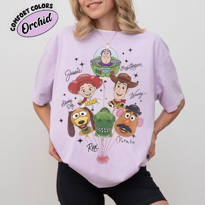 Puede incluir: Una camiseta de color lila claro con un gr&aacute;fico de los personajes de Toy Story: Buzz Lightyear, Woody, Jessie, Slinky Dog, Rex y Mr. Potato. La camiseta tiene el texto "Comfort Colors Orchid" en un dise&ntilde;o circular.
