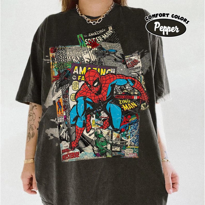 May include: Dark gray t-shirt featuring a colorful comic book design with the Amazing Spider-Man. The graphic includes the text "COMICS GROUP", "AMAZING SPIDER-MAN", and "ZING MAN". The shirt also has the text "COMFORT COLORS Pepper".