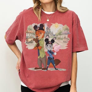 Puede incluir: Camiseta de tono rojo con una ilustraci&oacute;n de dibujos animados de Nick Wilde y Judy Hopps de Zootopia, con orejas de Mickey Mouse. El fondo muestra una escena de parque con un castillo y un carrito de comida. Los personajes sostienen algod&oacute;n de az&uacute;car.