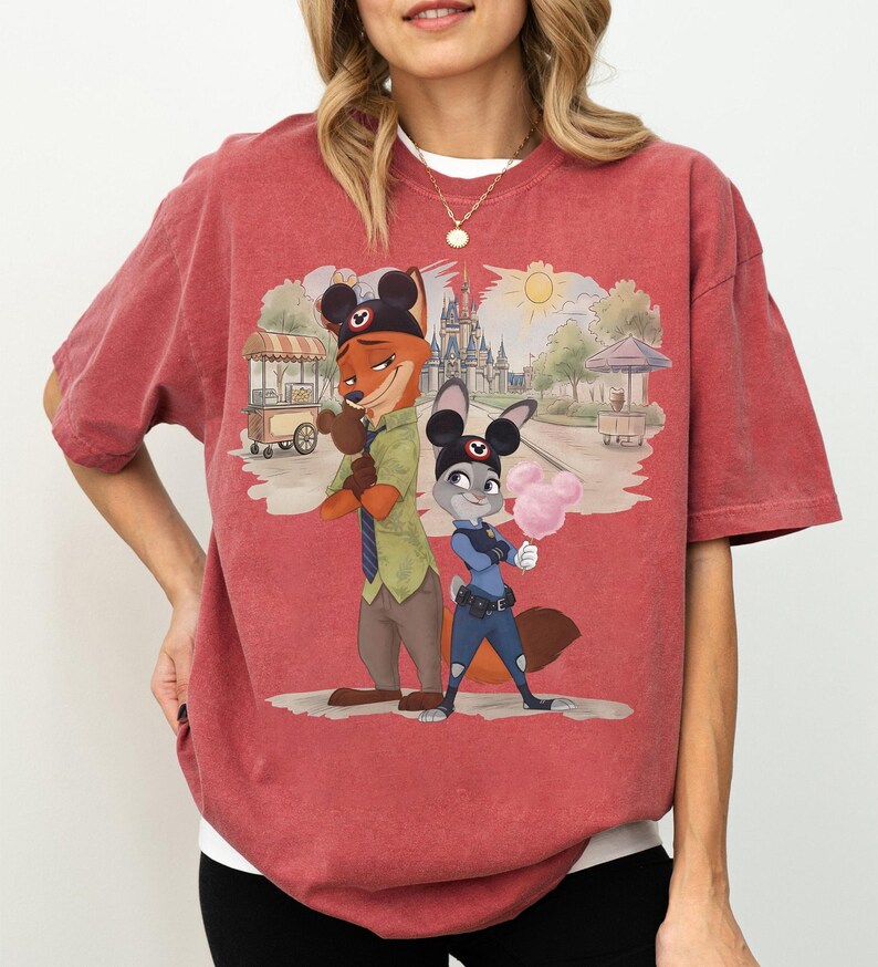 Puede incluir: Camiseta de tono rojo con una ilustraci&oacute;n de dibujos animados de Nick Wilde y Judy Hopps de Zootopia, con orejas de Mickey Mouse. El fondo muestra una escena de parque con un castillo y un carrito de comida. Los personajes sostienen algod&oacute;n de az&uacute;car.