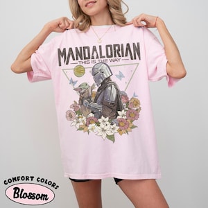May include: Light pink t-shirt with a graphic featuring The Mandalorian and Grogu surrounded by flowers and butterflies. The text "MANDALORIAN - THIS IS THE WAY" is printed across the top.