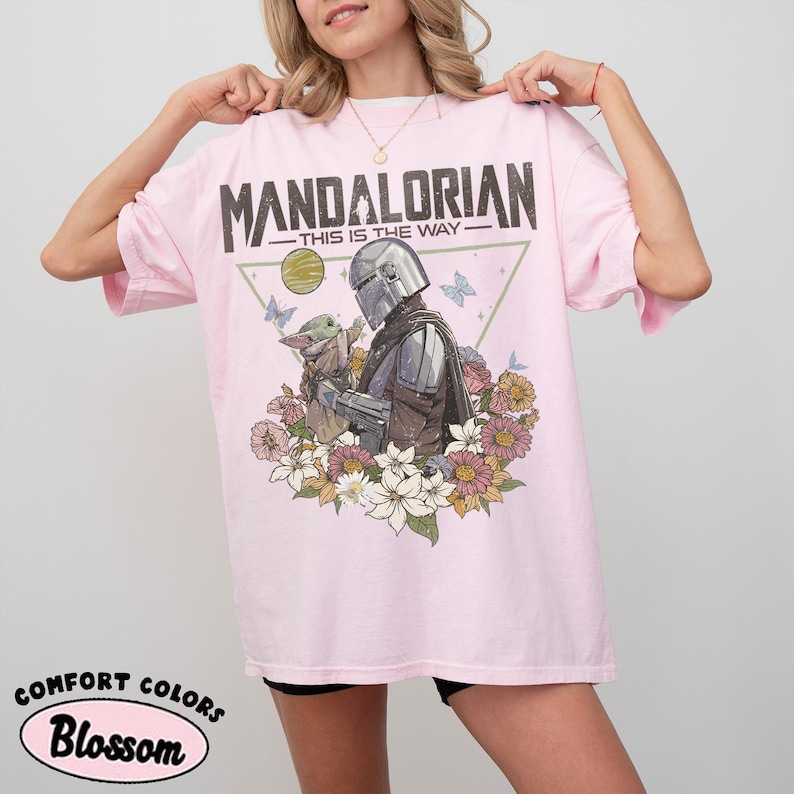 May include: Light pink t-shirt with a graphic featuring The Mandalorian and Grogu surrounded by flowers and butterflies. The text "MANDALORIAN - THIS IS THE WAY" is printed across the top.