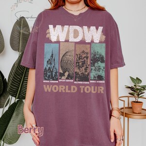 May include: A purple t-shirt with a graphic of the four theme parks at Walt Disney World. The text "WDW World Tour" is printed below the graphic. The parks are Magic Kingdom, Epcot Center, Hollywood Studios, and Animal Kingdom.