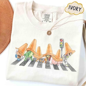 May include: Ivory-colored t-shirt featuring a whimsical illustration of cartoon characters crossing a crosswalk, each wearing an orange traffic cone. A traffic light and a gold necklace are also visible. The shirt is labeled "Comfort Colors."