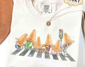 May include: Ivory-colored t-shirt featuring a whimsical illustration of cartoon characters crossing a crosswalk, each wearing an orange traffic cone. A traffic light and a gold necklace are also visible. The shirt is labeled "Comfort Colors."