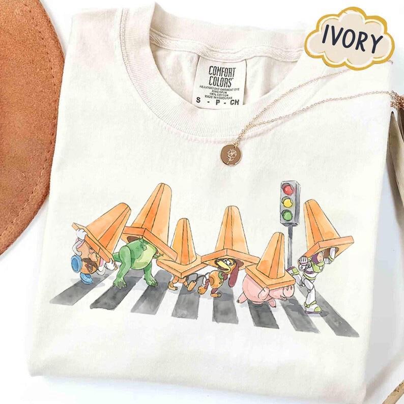 May include: Ivory-colored t-shirt featuring a whimsical illustration of cartoon characters crossing a crosswalk, each wearing an orange traffic cone. A traffic light and a gold necklace are also visible. The shirt is labeled "Comfort Colors."
