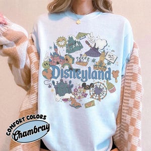 May include: Light blue sweatshirt with a Disneyland graphic. The graphic features various Disneyland icons, including the castle, a Ferris wheel, and the word "Disneyland" in a whimsical font. The sweatshirt has the words "Comfort Colors" and "Chambray" printed on it.