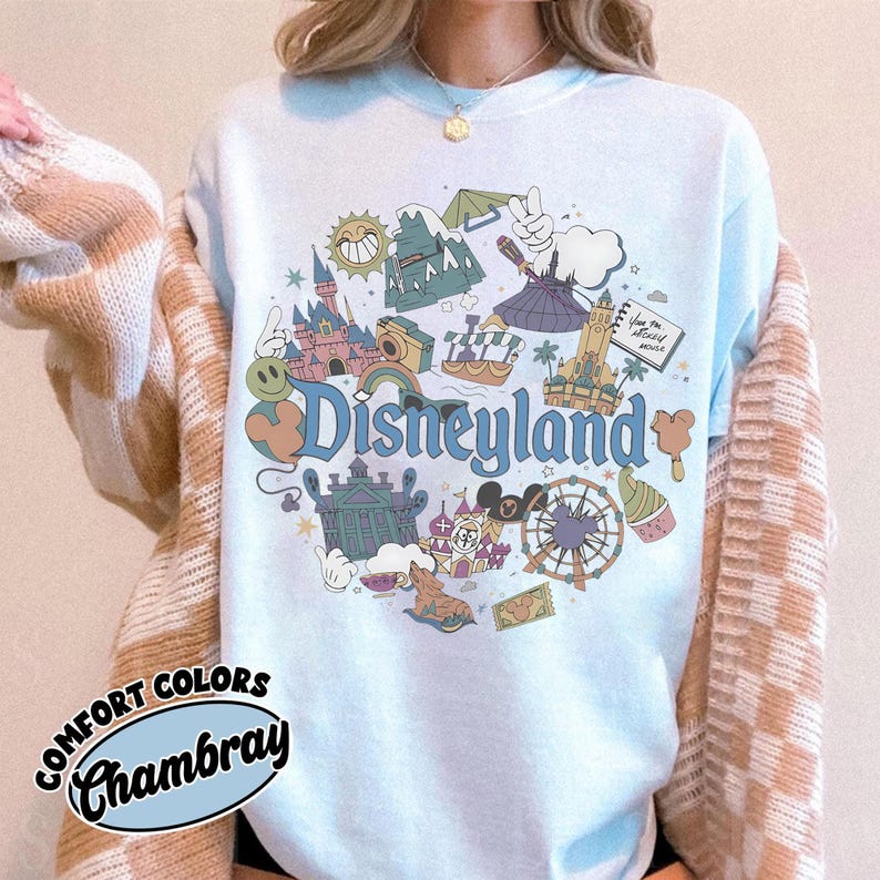 May include: Light blue sweatshirt with a Disneyland graphic. The graphic features various Disneyland icons, including the castle, a Ferris wheel, and the word "Disneyland" in a whimsical font. The sweatshirt has the words "Comfort Colors" and "Chambray" printed on it.