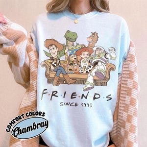 May include: A light blue crewneck sweatshirt featuring a graphic print of Toy Story characters sitting on a couch. The text below the characters reads FRIENDS SINCE 1995. The shirt is layered under a beige and white checkered cardigan sweater.