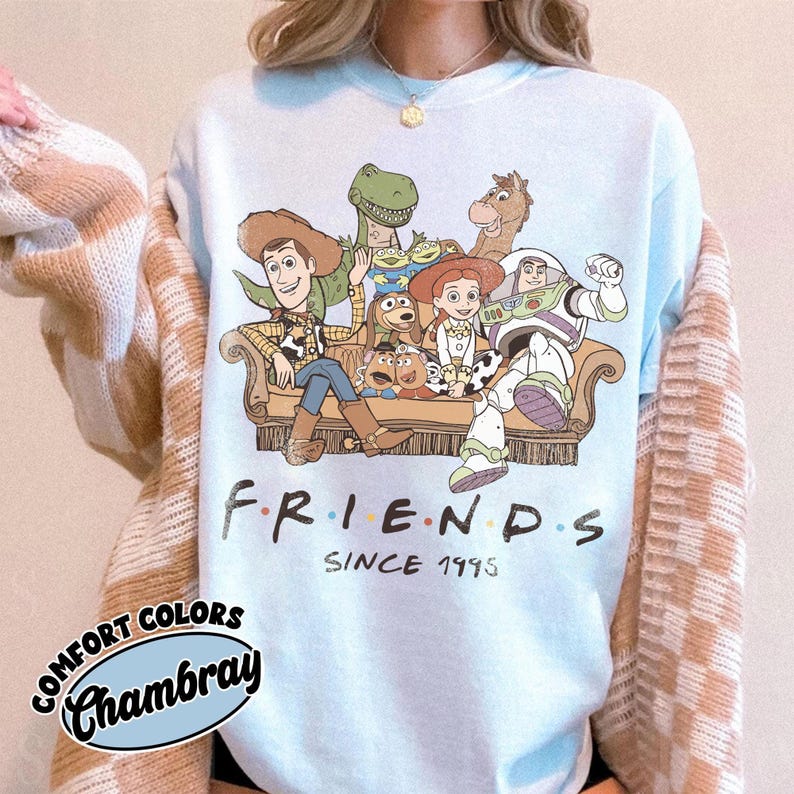 May include: A light blue crewneck sweatshirt featuring a graphic print of Toy Story characters sitting on a couch. The text below the characters reads FRIENDS SINCE 1995. The shirt is layered under a beige and white checkered cardigan sweater.
